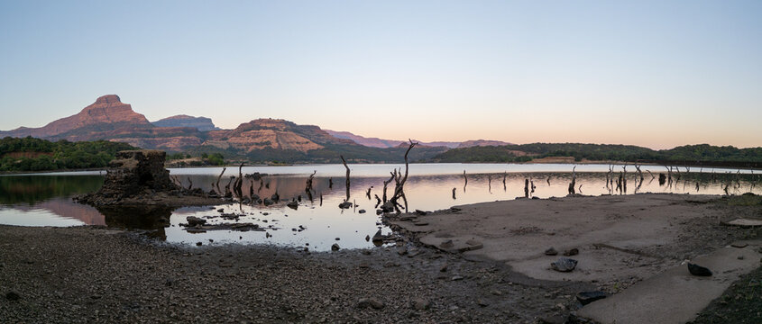 Wide panoramic view of Ghatghar Lake in Bhandardara, showing dry shoreline, calm water, mountain silhouettes and tree stumps at sunset. A dramatic natural landscape scene in Maharashtra.