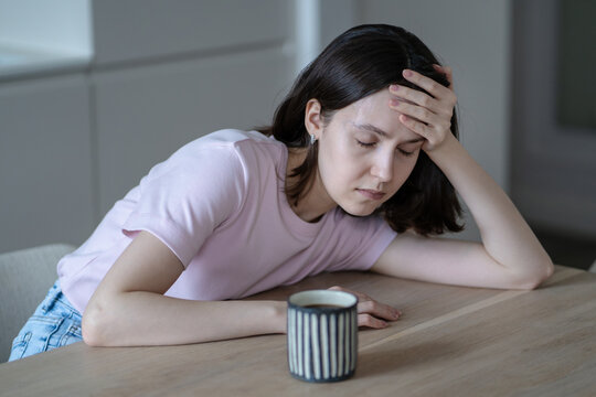 Exhausted young woman sits at table in morning with cup of coffee, resting head on hand with closed eyes, shows signs of morning fatigue and apathy. Sleepy female sits in kitchen after restless night.