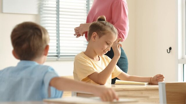 Beautiful teacher explaining test while caucasian boy doing classwork and listening explanation. Cute child looking at teacher while taking a notes or writing answer in paper at classroom. Pedagogy.