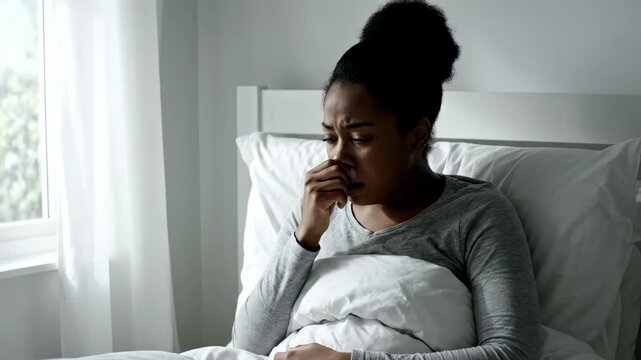 Unwell young black woman sitting up in bed and suffering from a cold or flu next to a bright window, concept for health crisis reports, medical advertising and mental illness issues