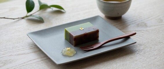 Delicate confection on a minimalist gray plate, featuring a delectable cake adorned with golden accents and set against a backdrop that includes a cup of tea, a wooden spoon, and a sprig of greenery.