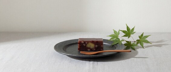 A Japanese sweet treat, wagashi, exquisitely presented on a minimalist grey plate, hinting at flavors and textures, with fresh maple leaf adding a touch of nature's beauty. 