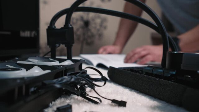 A person in the background flips through a manual while CPU cooler fans, cables, and water block from a liquid cooling system lie on the foreground
