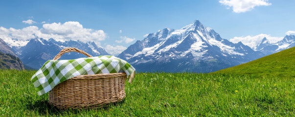 picnic basket sits on lush green grass, framed by majestic snow-capped mountains under a bright blue sky.