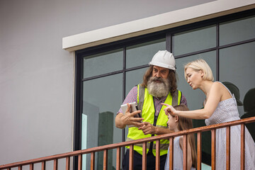 Experienced bearded foreman shows new home plans on a tablet to a smiling mother and daughter over terrace, highlighting smart construction technology and a warm, hopeful future in their house