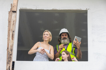 Experienced bearded foreman shows new home plans on a tablet to a smiling mother and daughter over terrace, highlighting smart construction technology and a warm, hopeful future in their house