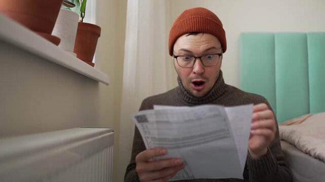 Surprised man wearing warm clothes inside his home becoming very emotional and upset after seeing the high cost of his utility bills, frantically checking his cold radiator