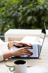 Close-up of a woman&rsquo;s hands typing on a laptop at an outdoor workspace with a coffee cup on the table