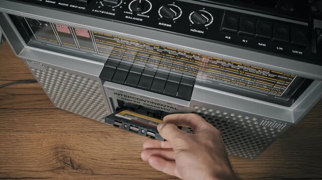 Top-down view of male hands inserting a retro audio cassette into a silver boombox. The hands press the black start button to play music on a wooden table surface.