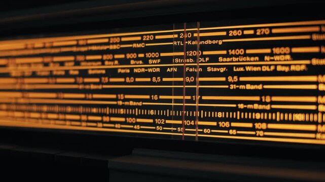 Extreme close-up of a tuning needle moving across an illuminated radio dial. The vintage receiver glows warm orange as it searches for stations on the analog scale.