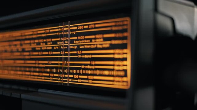 Side-angle view of a hand turning a knob to tune a vintage radio receiver. The needle moves across the illuminated orange frequency dial searching for broadcast stations.