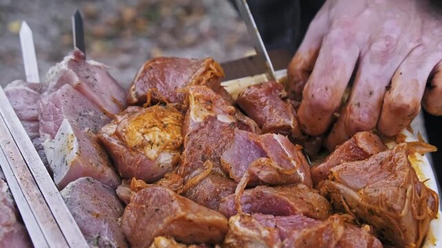 Male hands mix and cut pieces of raw spiced meat for shish kebabs outdoors. The preparation involves seasoning pork chunks before placing them on metal skewers for grilling.