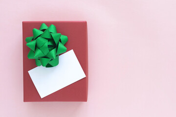 A beautiful holiday present for giving and advertising concepts with copy space. Flat lay top view of an elegant red gift box with green bow and a blank white card for custom text on pink paper.