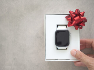 Modern smartwatch in a white gift box with a red ribbon bow held by a hand. Perfect for tech gift concepts, Christmas, or birthdays on a grey textured background with copy space for your text.
