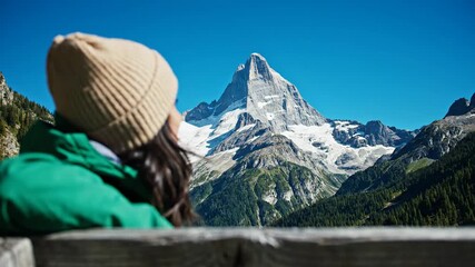 Young woman in a beanie gazes at a majestic snow-capped mountain peak in a scenic alpine landscape.