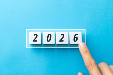 Minimalist concept of transition to 2026 with white cubes on a blue background. Features loading bar, finger touching block, and flipping 2025 to 2026 to represent future goals and business planning.