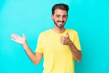 Fototapeta premium Young caucasian man isolated on blue background holding copyspace imaginary on the palm to insert an ad and with thumbs up