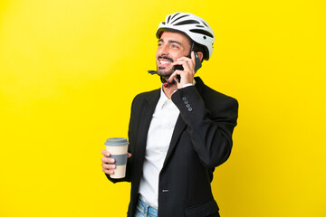 Business caucasian man with a bike helmet isolated on yellow background holding coffee to take away and a mobile