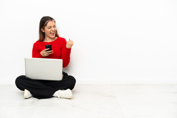 Young caucasian woman with a laptop sitting on the floor with phone in victory position