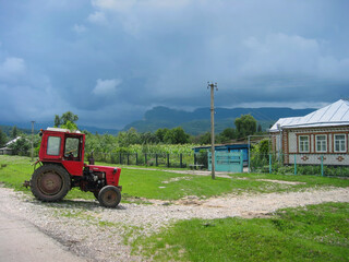 A summer rural landscape with a red tractor and a one-story brick house. Mountains are visible in the distance. The sky is overcast with dark clouds. Akhmetovskaya village, Krasnodar Krai, Russia.