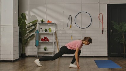 A woman in a pink shirt and black leggings performs a lunge stretch on a wooden floor in a gym room. Exercise, fitness training for women, healthy lifestyle.