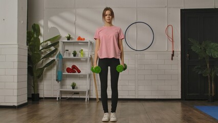 A young woman in a pink shirt and black pants is standing in a gym setting holding green dumbbells. Exercise, fitness training for women, healthy lifestyle.