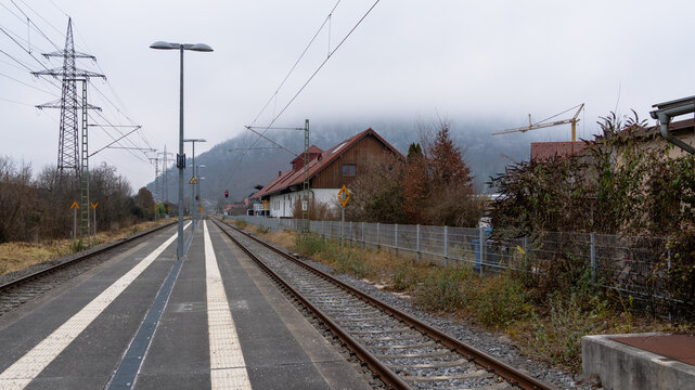 A wide-angle view of a small rural train station in Freilassing, Bayern, Germany, with misty hills in the background, empty platform, railway tracks, and power lines under cloudy winter conditions.