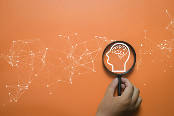Hand holding magnifying glass focusing on head brain icon with digital network lines on orange background. Concept of artificial intelligence, mental health check,creative idea,and machine learning.