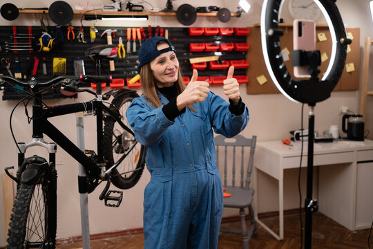 female worker blogger influencer filming video mobile phone make tutorial stream blog showing thumbs-up while repairing bicycle in Bike workshop interior.