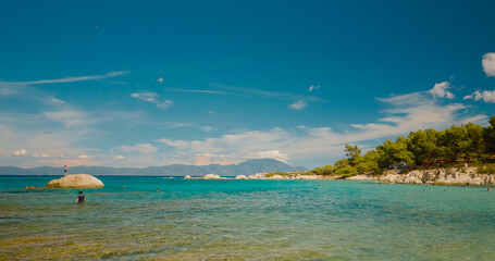 Sunbathers and swimmers relish the warm sun and crystal clear waters on a scenic Greek beach, surrounded by rocky shores and lush greenery on a summer day.