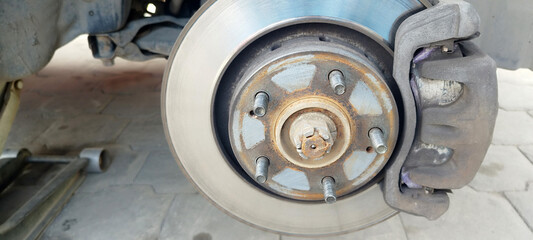 A close-up of a vehicle&rsquo;s wheel hub assembly, showing a brake rotor, rusted lug studs, the brake caliper, and the center axle nut, likely undergoing maintenance