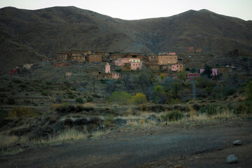 A remote Berber village clings to the slopes of the Atlas Mountains, Morocco, with earth-toned houses scattered across a dry valley under muted evening light.