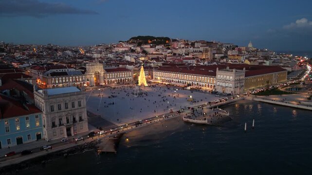Cinematic aerial view of Praca do Comercio in Lisbon Portugal at blue hour festive Christmas tree lights glowing across the main square, riverfront buildings creating a holiday city mood
