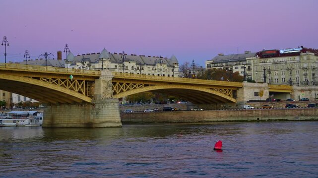 Side view of the historical yellow Margaret Bridge across the Danube river in Budapest, Hungary, during a soft purple sunset with city buildings in the background
