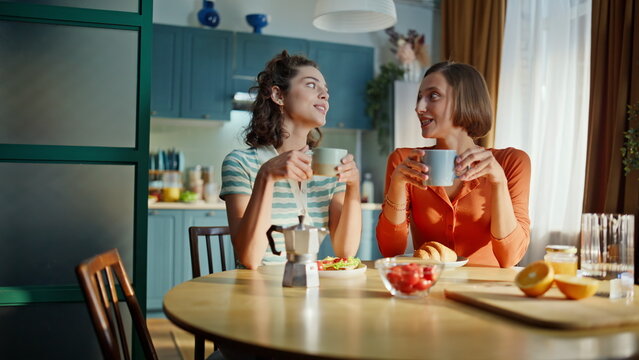 Two besties holding cups with hot tea sitting together at apartment kitchen. 
