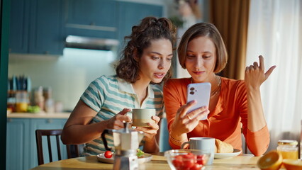 Cheerful besties shopping online browsing cellphone app at home kitchen closeup.