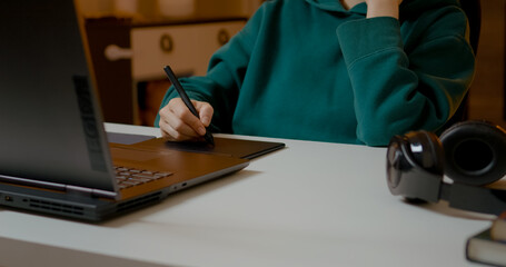 Graphic designer focusing intently on digital drawing, using tablet and laptop in home workspace with creative concentration