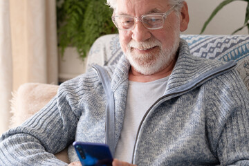Elderly 70s man seated on sofa browsing on smartphone, modern senior male looking at cell screen surfing the net or social media, older generation and modern tech application easy usage concept