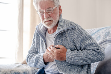A senior man over 70 wearing a blue sweater sitting on the couch at home suffers from severe chest pain - heart disease and high blood pressure in the elderly