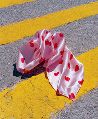 Pink heart silk scarf lying on asphalt beside bold yellow street lines, romantic textile lost on road, valentine detail in urban sunlight