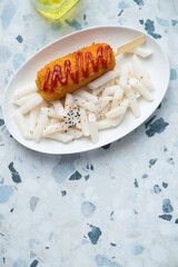 Plate with korean tteokbokki or rice cakes and a corn-dog on a white and blue granite background, vertical shot with space, top view