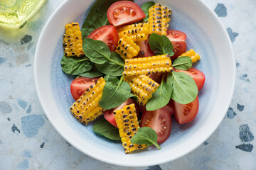 Plate of salad with tomato wedges, grilled corn and fresh spinach, horizontal shot on a white and blue granite surface, middle close-up