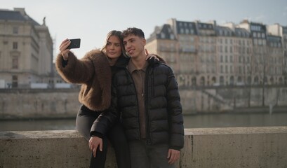A romantic couple is taking a selfie with a smartphone in Paris France with the Seine River and buildings in the background on a sunny day. This image is suitable for travel and tourism related conten