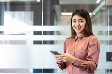 Young businesswoman smiling cheerfully and engaging with her smartphone in a modern corporate office environment, representing professional communication, technology use, and happiness at work