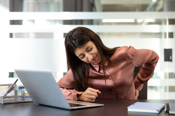 Young woman feeling intense back pain and discomfort while sitting at her office desk, highlighting...