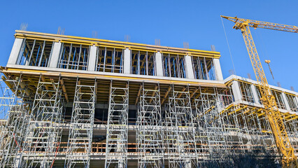 New building under construction, featuring a multi story concrete structure with extensive scaffolding and formwork, supported by a large yellow tower crane under a clear blue sky