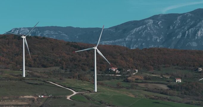 Aerial view of wind turbines standing tall on a verdant hillside with houses, reflecting the clear blue sky, creating a striking contrast, Serre, Campania, Italy.
