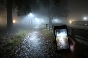 Hand holding smartphone capturing a mystical foggy path with dim streetlights at night