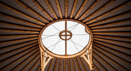 Interior low-angle shot of a traditional yurt ceiling featuring radial wooden beams and a central circular skylight (toono) against a bright sky