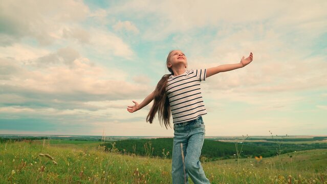 Kid girl plays on lawn under sky, childhood dream travel. Child girl enjoys green nature in summer, raising her arms. Kid in park. Slow motion. Little girl plays in meadow, dream of fly. Happy family